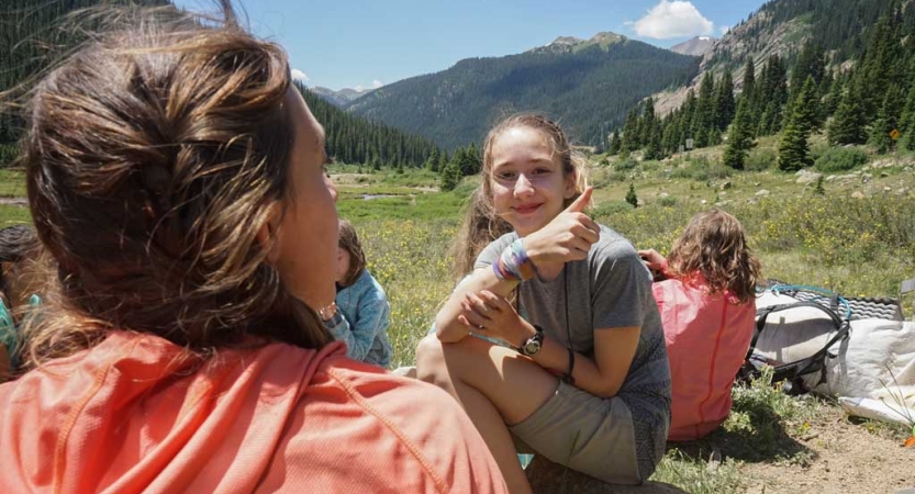 A group sits in a green mountain valley. One of them smiles at the camera and gives a thumbs up. 